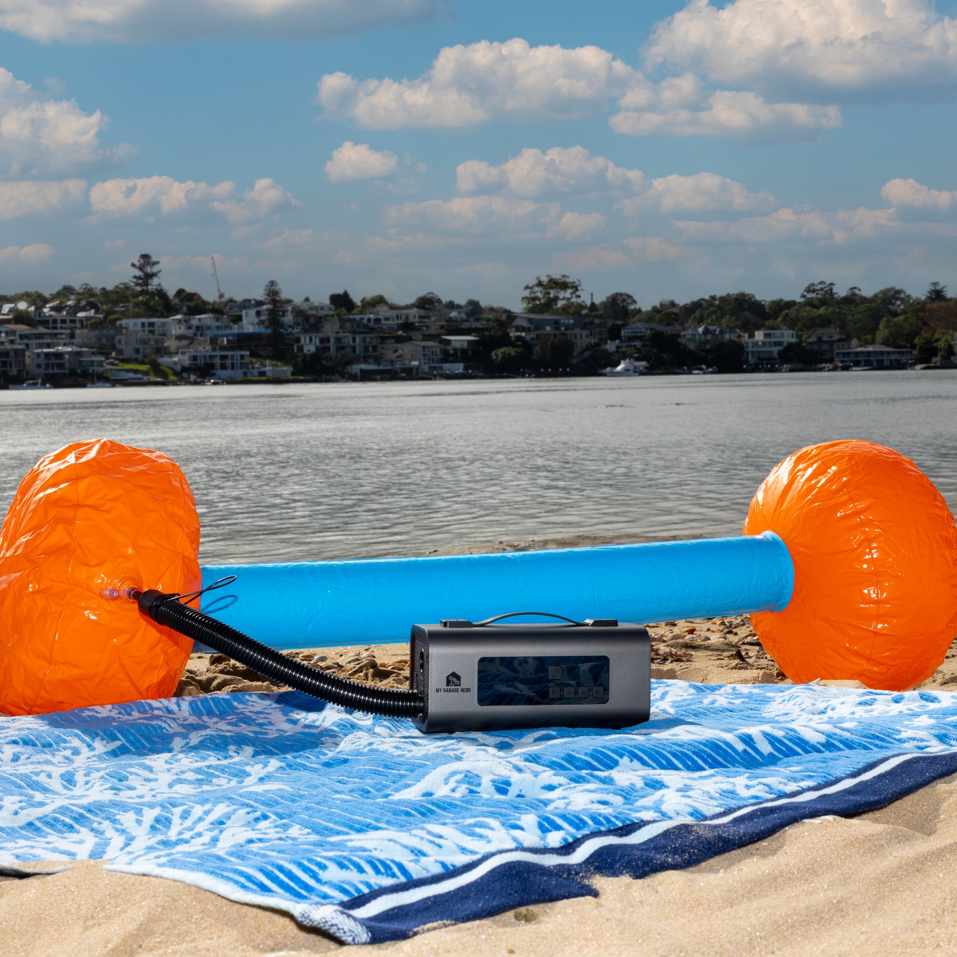 Boost N’ Inflate inflating an orange and blue inflatable dumbbell on a beach towel by the water, with houses and trees visible across the shoreline under a partly cloudy sky.