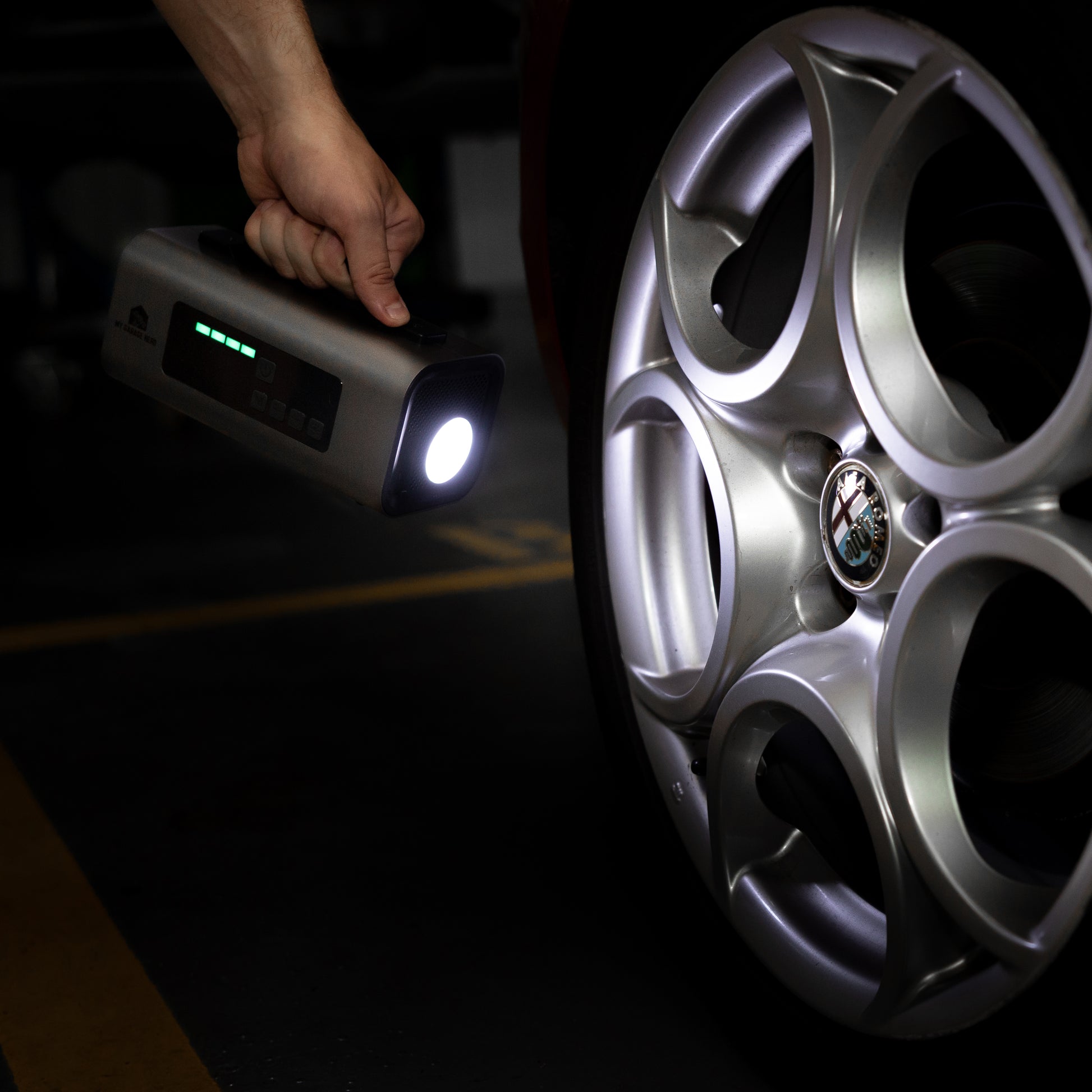 Person holding the Boost N’ Inflate with its LED light turned on, illuminating an Alfa Romeo alloy wheel in a dimly lit garage.
