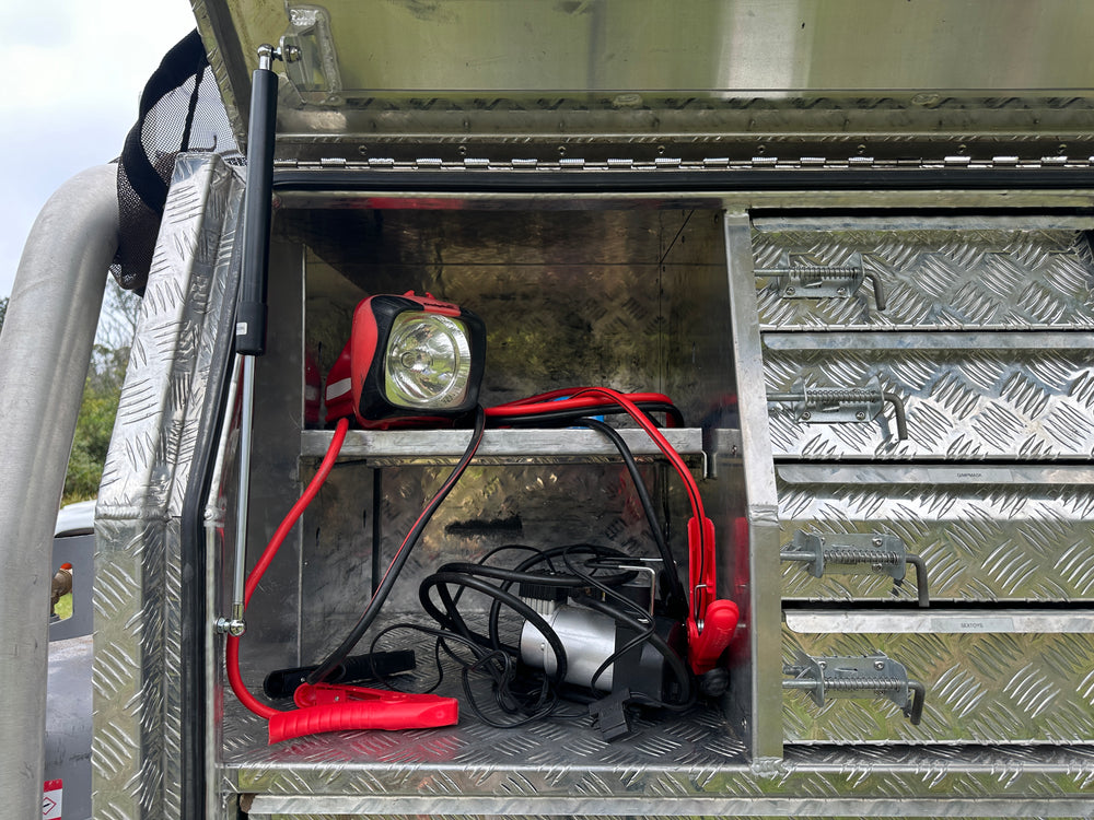 Ute tray storage compartment with red and black jumper cables, air compressor, and a torch taking up significant space inside an aluminium checker plate toolbox with multiple drawers.