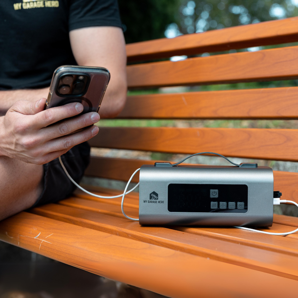 A portable jumpstarter and tyre inflator, the "boost n inflate", sitting on a park bench facing directly towards the camera. it has an iphone cable plugged into its port and is charging an iphone 13 pro