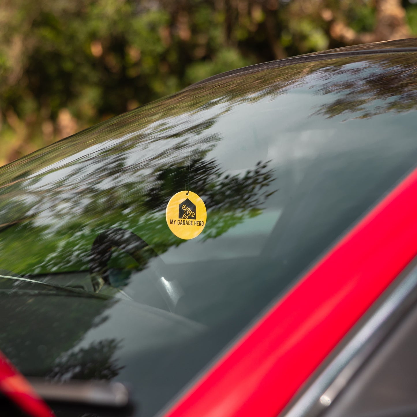 My Garage Hero circular air freshener hanging from a rear-view mirror, shown inside a car with trees and sky reflecting on the windscreen for a natural outdoor lifestyle look.