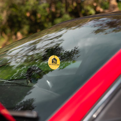 My Garage Hero circular air freshener hanging from a rear-view mirror, shown inside a car with trees and sky reflecting on the windscreen for a natural outdoor lifestyle look.