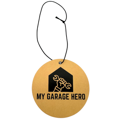 A circular air freshener with a brown background and black text that reads 'MY GARAGE HERO', designed with a graphic of a wrench and a house silhouette. A black string is attached for hanging.