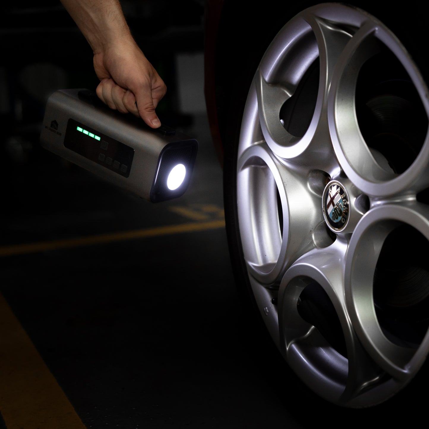 Person holding the Boost N’ Inflate with its LED torch switched on, illuminating an Alfa Romeo alloy wheel inside a dimly lit garage.