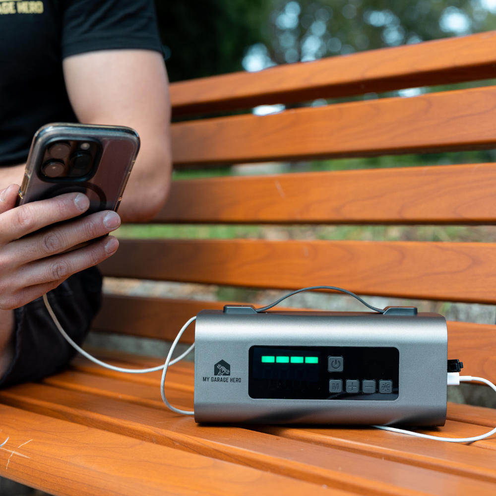 Person sitting on a wooden park bench charging their smartphone with the Boost N’ Inflate, which is being used as a portable power bank in an outdoor setting.