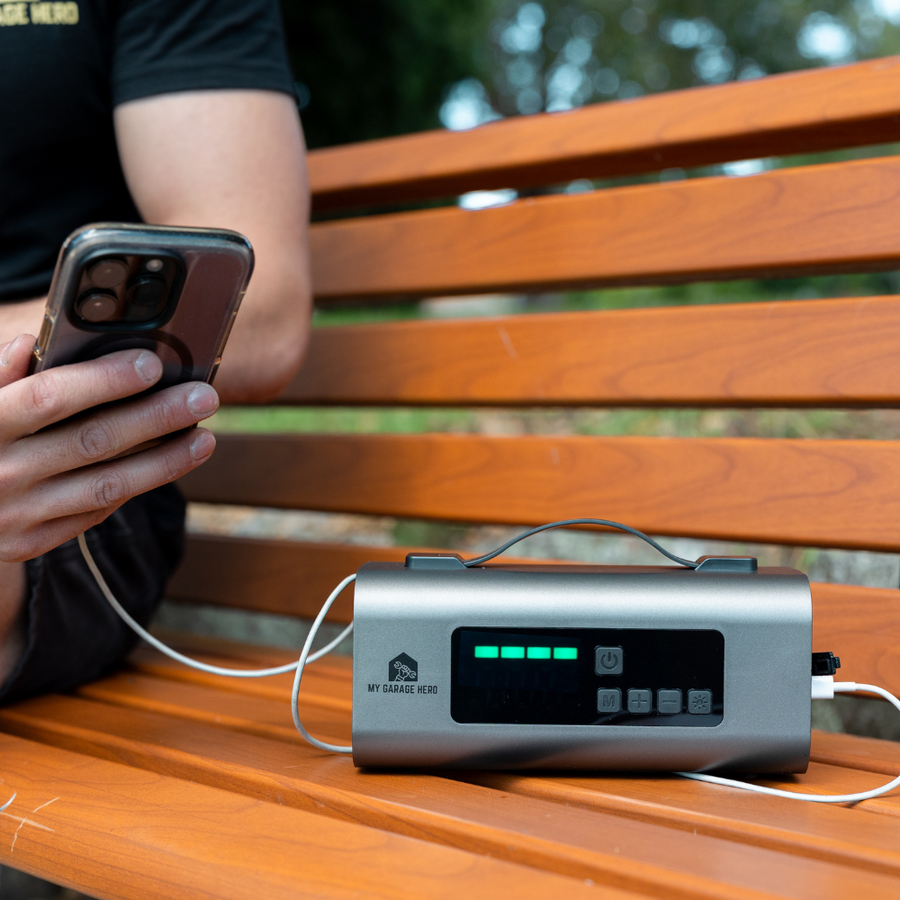 Person sitting on a wooden park bench charging their smartphone with the Boost N’ Inflate, which is being used as a portable power bank in an outdoor setting.