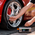 Person inflating a car tyre using the Boost N’ Inflate portable jump starter and tyre inflator, with the unit resting on the ground beside an Alfa Romeo alloy wheel during real-world use.
