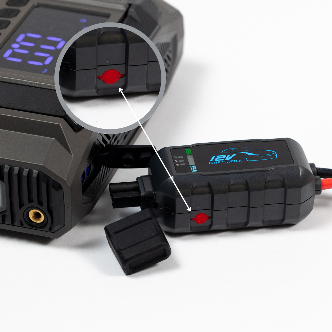Close-up of a jump starter with digital display on a white background, with a zoom in lens and arrow display a red button on the side of the jump start cables. 